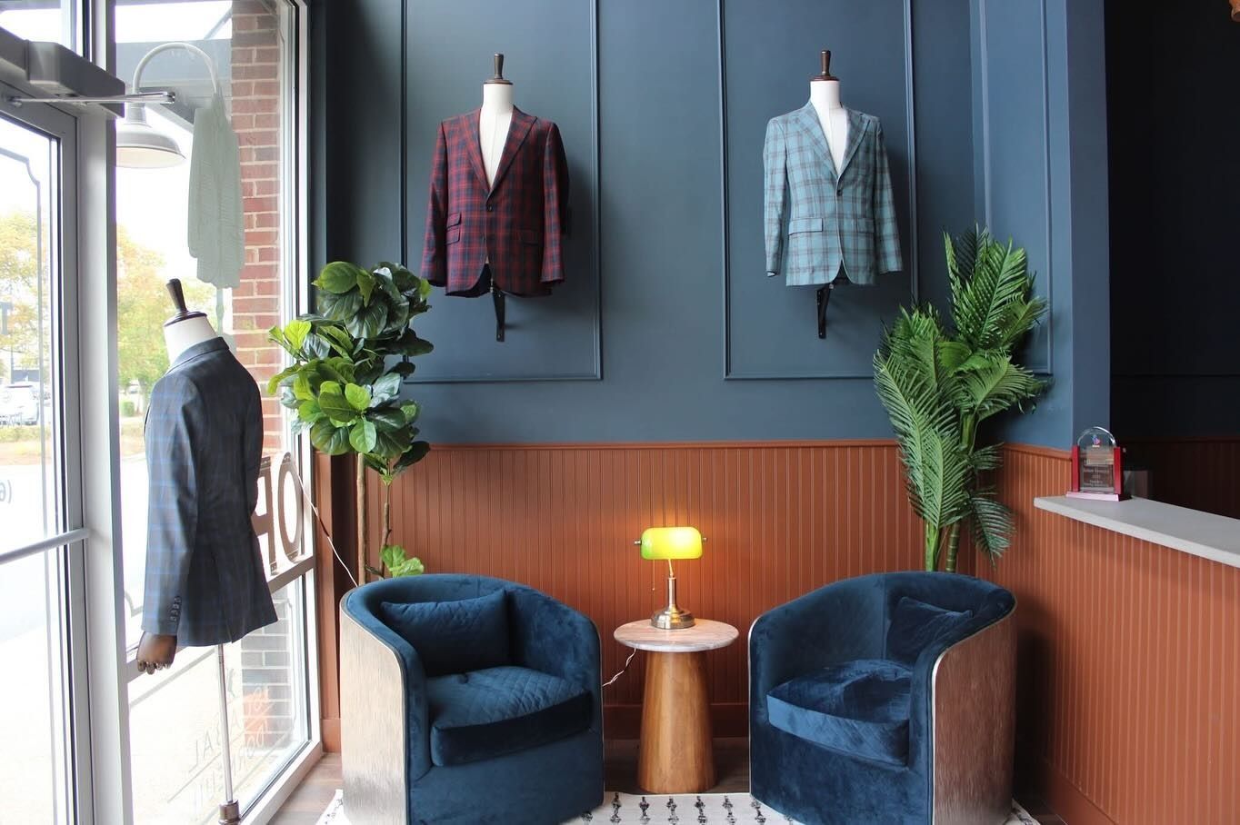 Tailor shop interior with blue velvet armchairs, jackets on display, and plants.