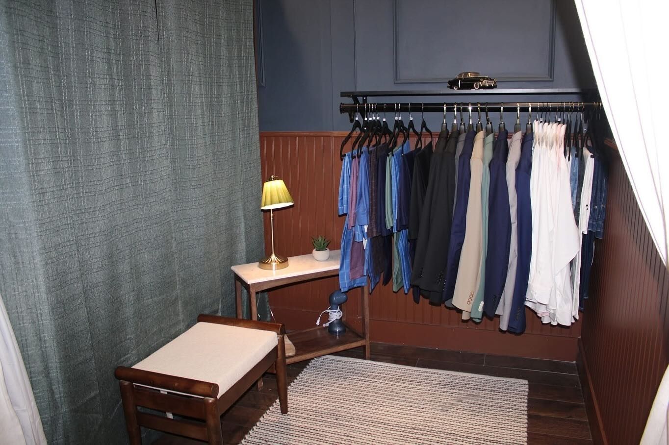 Clothing rack with garments in a small room with a bench, table with lamp, and green curtain.