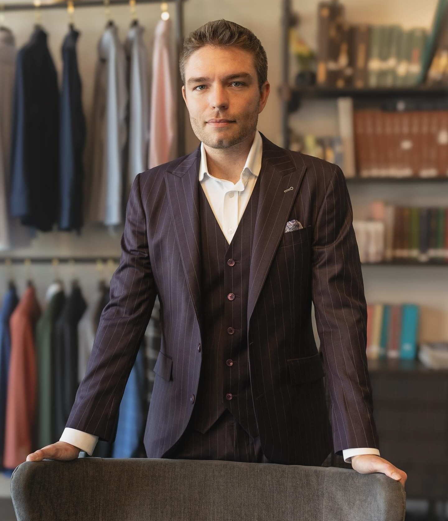 Man in a suit leans on a bar, looking directly at the camera. Outdoor setting, neutral colors.
