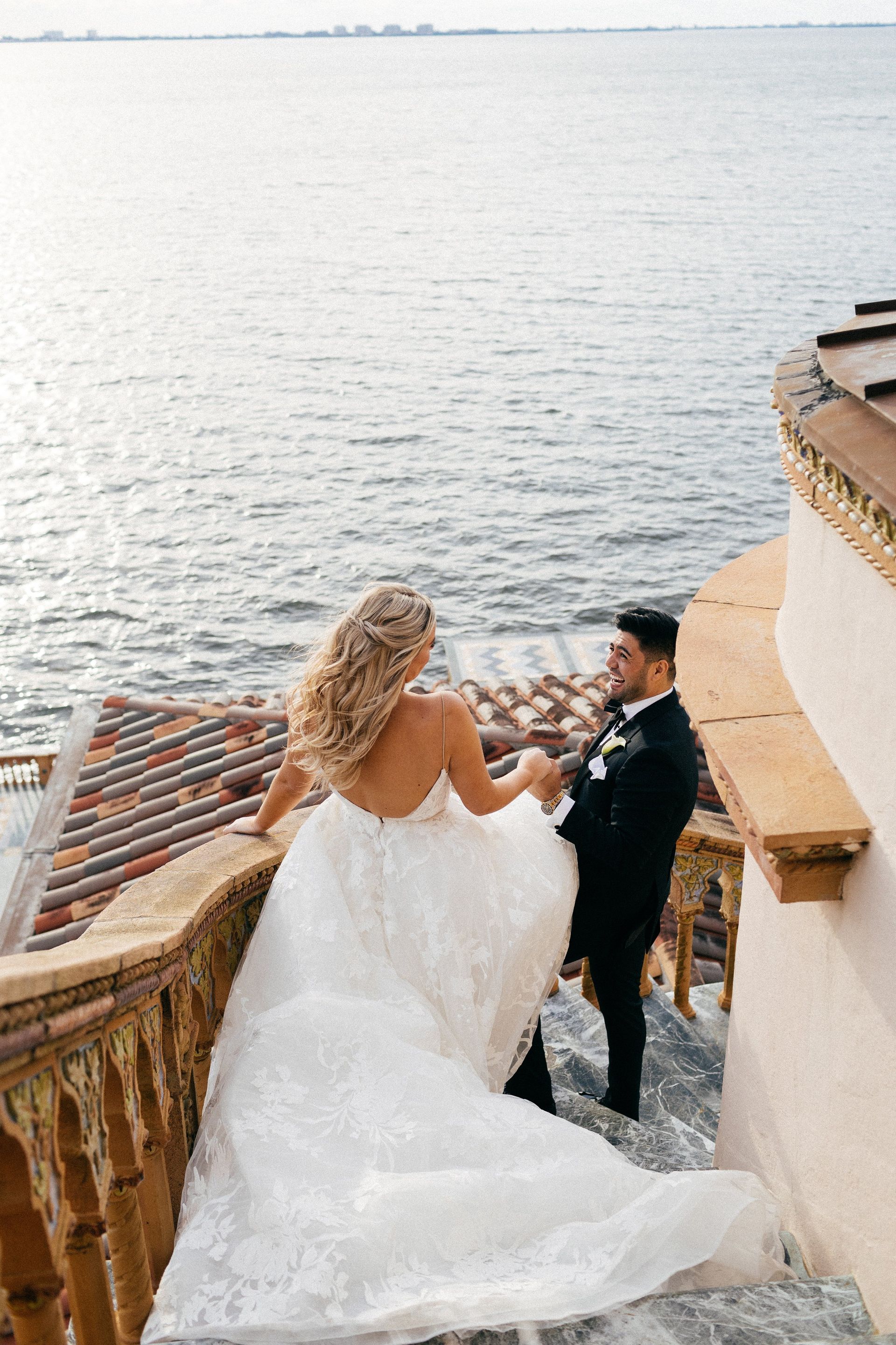 A bride and groom are standing on a balcony overlooking the ocean.