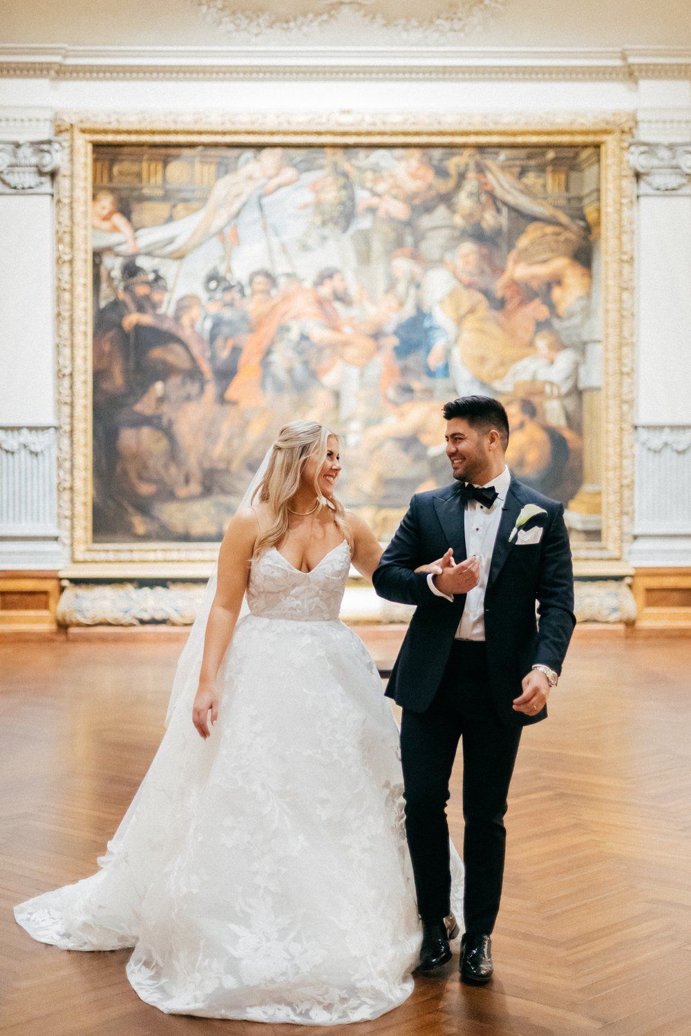 A bride and groom are standing in front of a large painting.