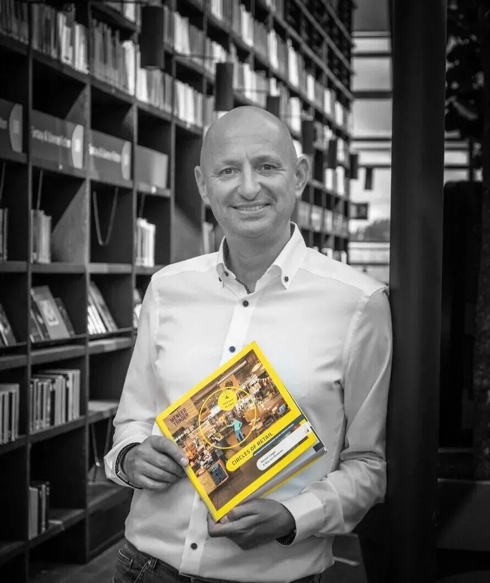 Een glimlachend persoon in een wit shirt staat in een bibliotheek en houdt een geel boek naar de camera gericht.