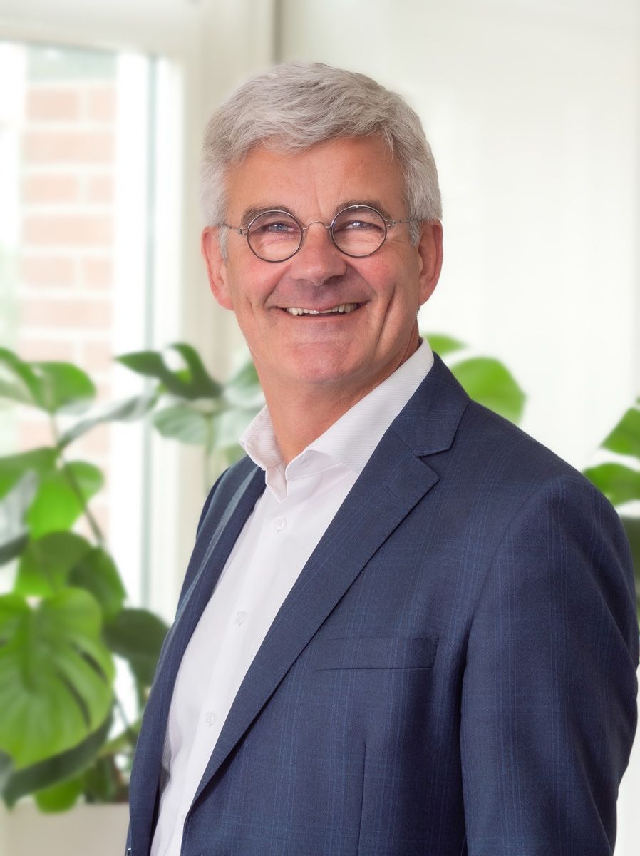 Een man in pak en bril lacht voor een raam