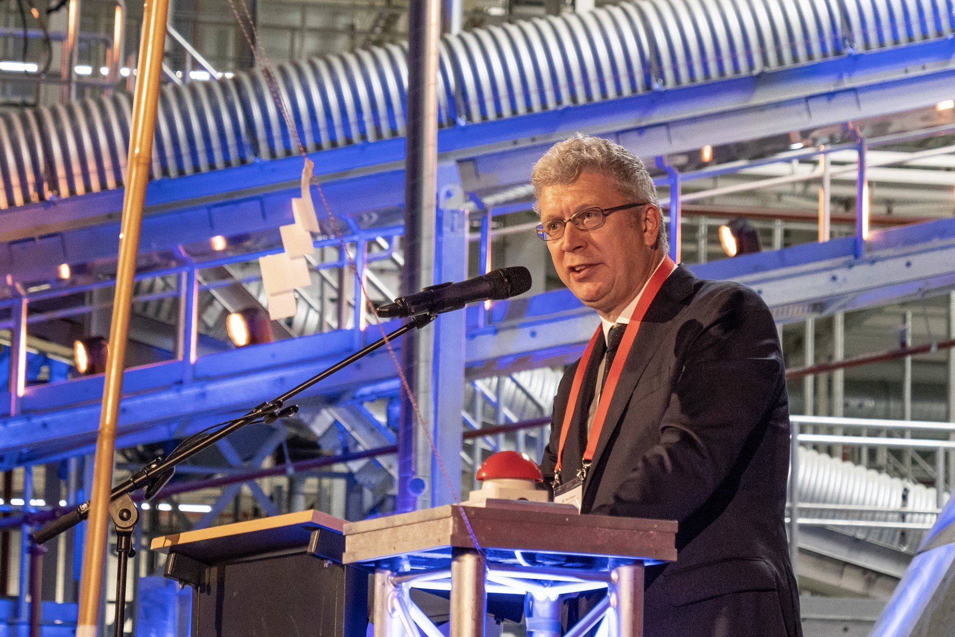 Een man in pak en stropdas staat op een podium en spreekt in een microfoon.