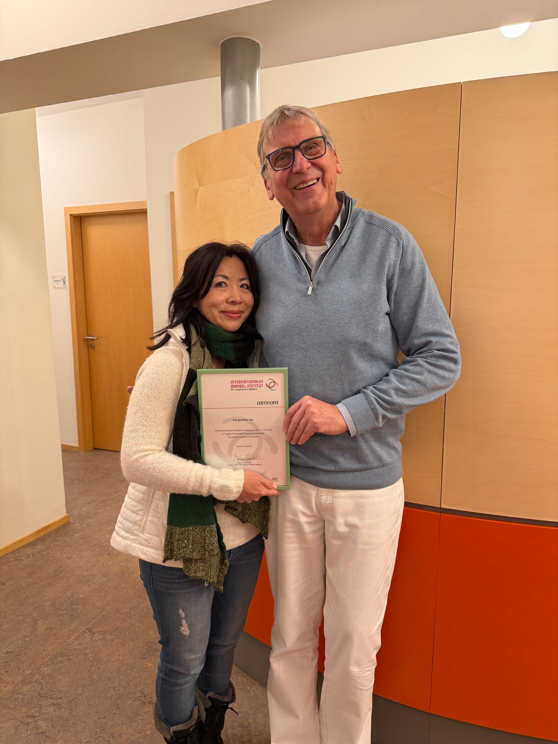 Woman and man smiling, holding certificate. They are in a building with neutral-colored walls.