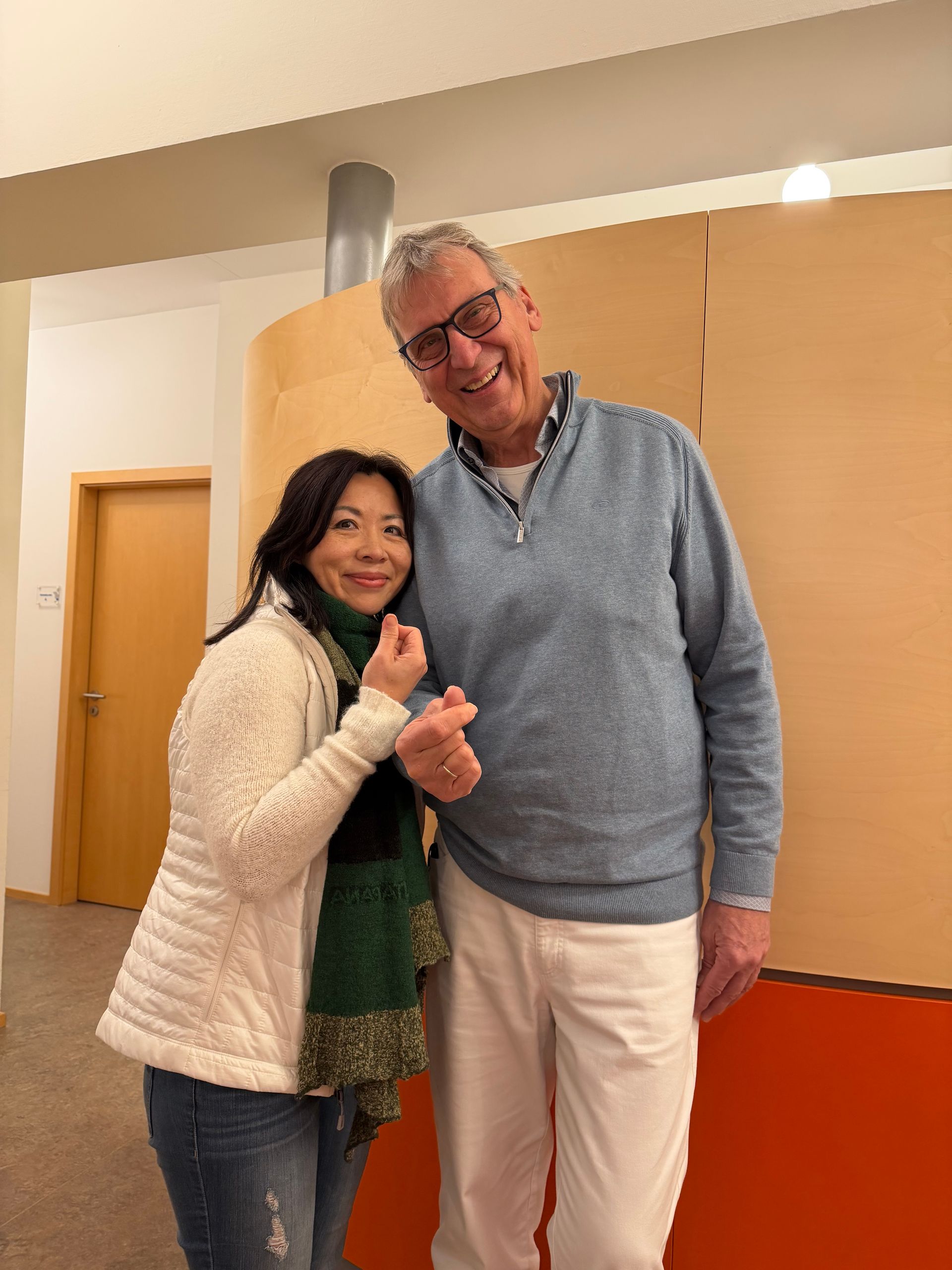 Woman and man smiling, holding hands. She wears a white jacket, green scarf, jeans. He wears a blue sweater, white pants. Interior setting.