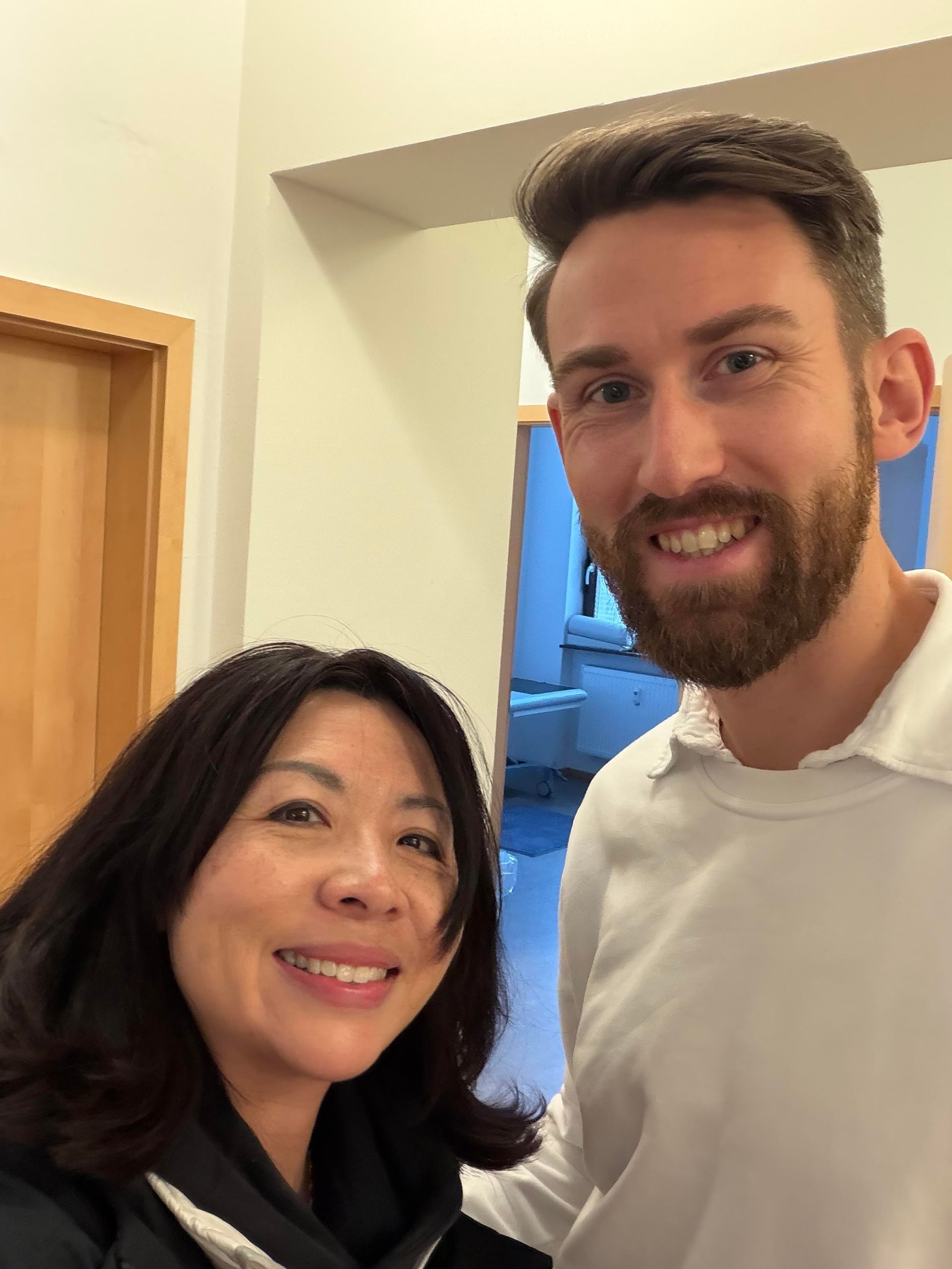 Woman and man smiling, taking a selfie. Man has a beard, wearing white shirt. Interior setting.