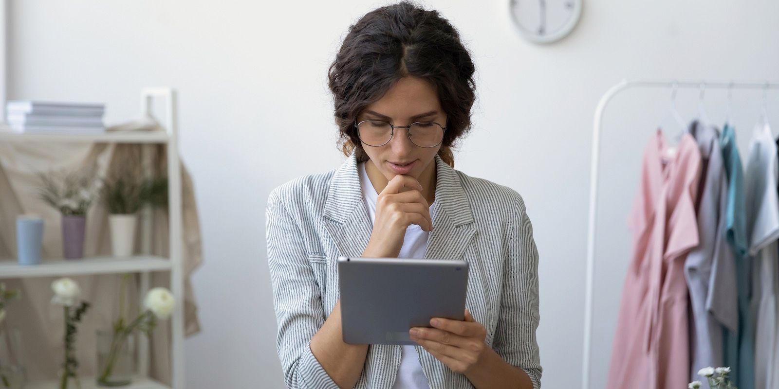 Woman in glasses looks at a tablet, hand on chin. Clothes rack and shelf in background.