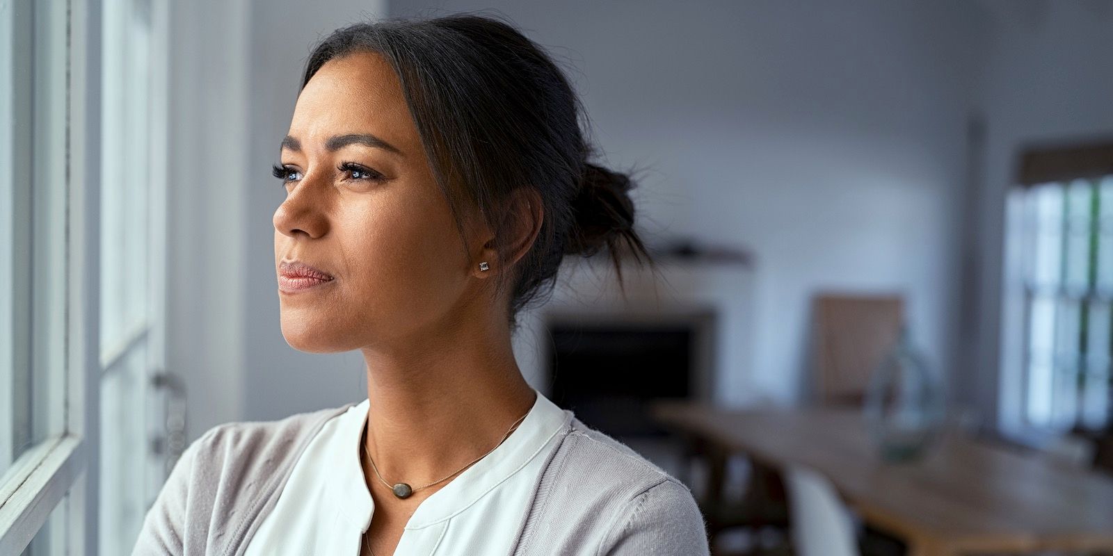 Woman gazing out a window, thoughtful expression. Soft lighting, neutral background with blurred interior.