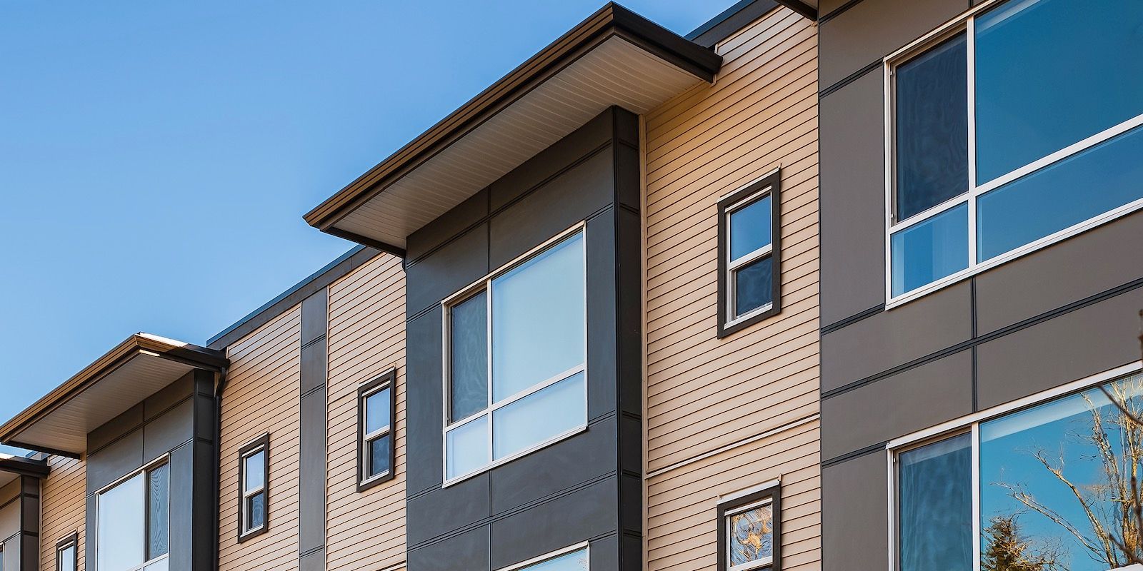 Modern building exterior with gray and beige siding, large windows, and a blue sky backdrop.