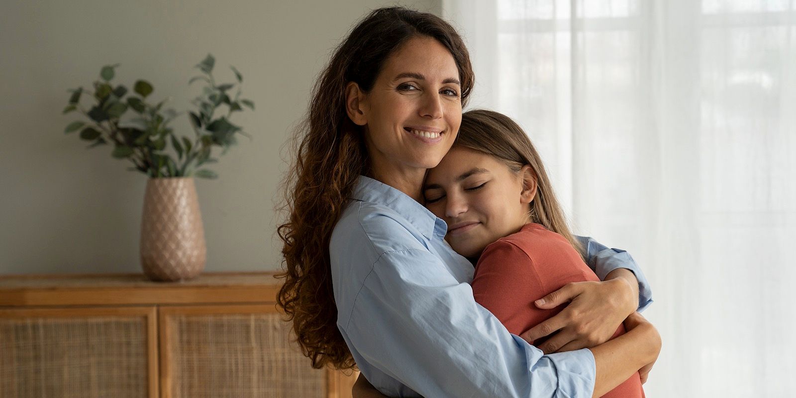 Woman hugs girl, both smiling, in front of a window and cabinet with a vase of greenery.