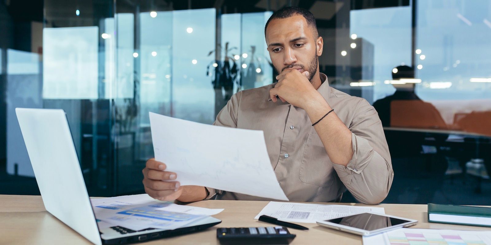 Man at a desk in an office, reviewing papers with a laptop and tablet, looking concerned.