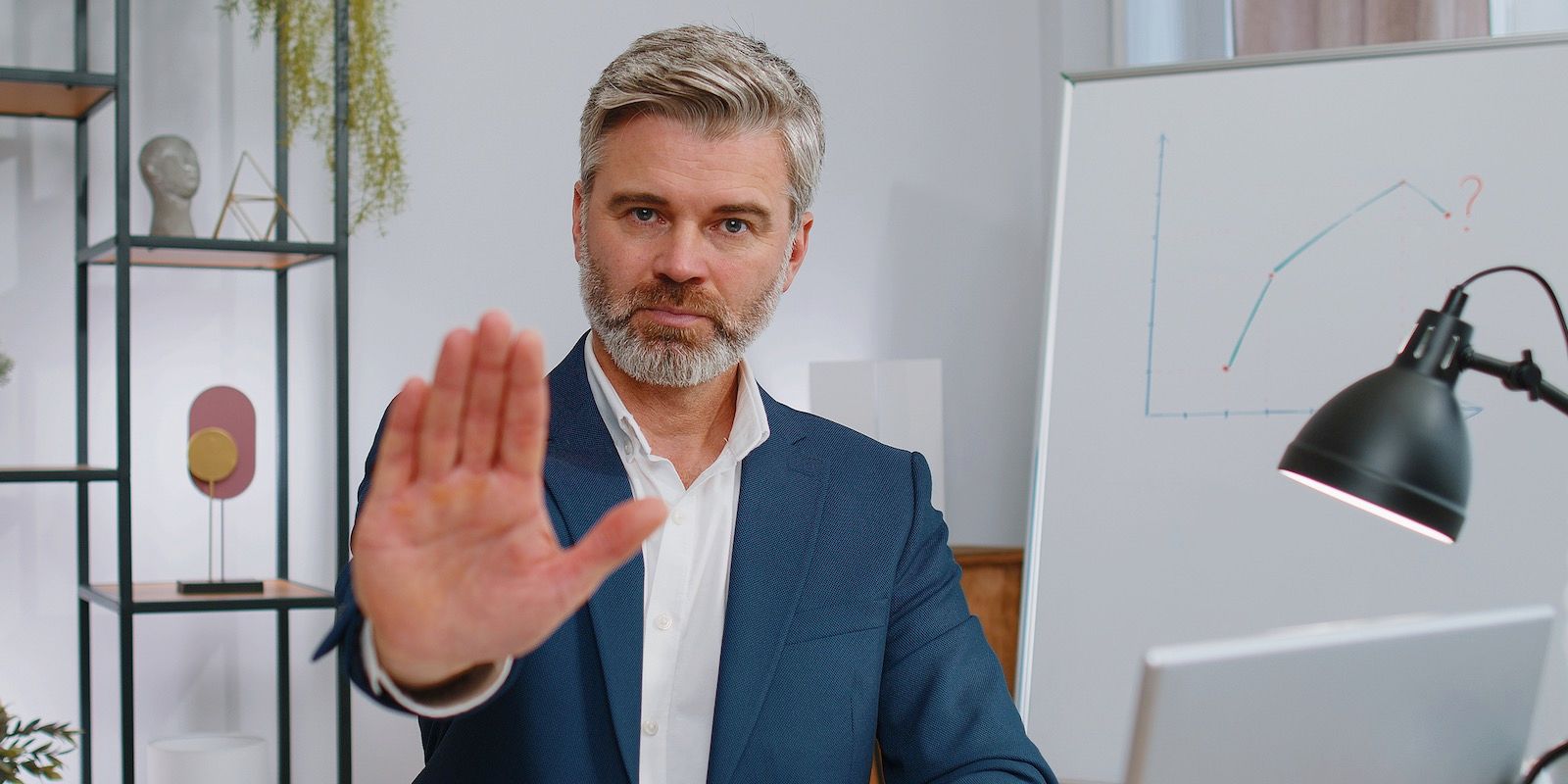 Man in suit gestures stop with open palm, indoors, desk with lamp and whiteboard in background.