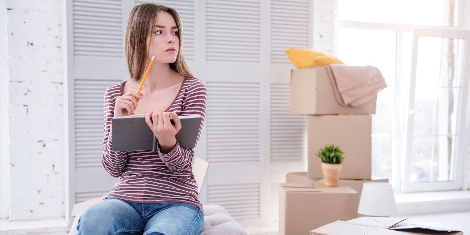 Woman with notebook and pencil, near moving boxes, looking thoughtful.