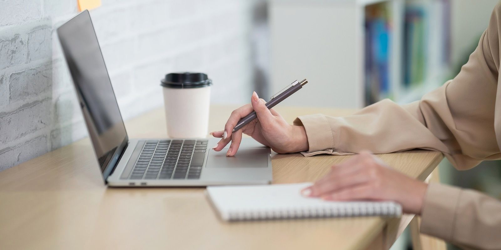 Person using laptop, holding pen, and writing in notebook on desk with coffee.