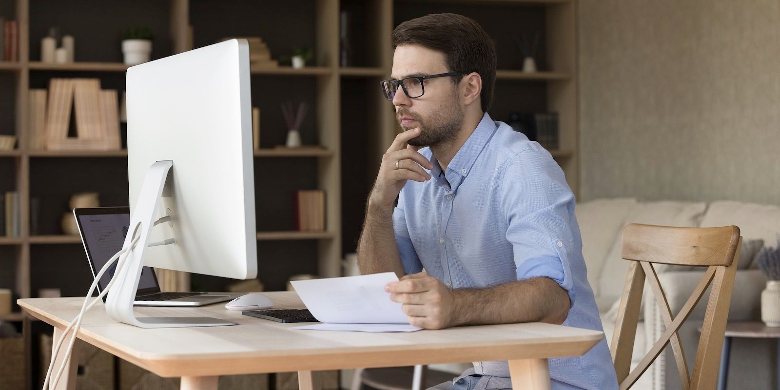 Man wearing glasses at desk, looking at computer screen, holding papers, in home office.