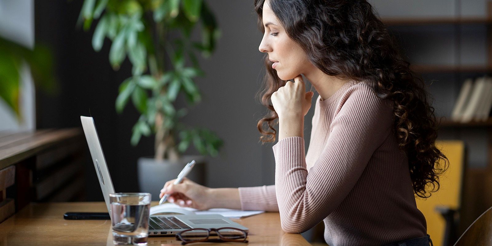 Woman working on a laptop at a desk, with pen and paper, glass of water, and a plant in the background.