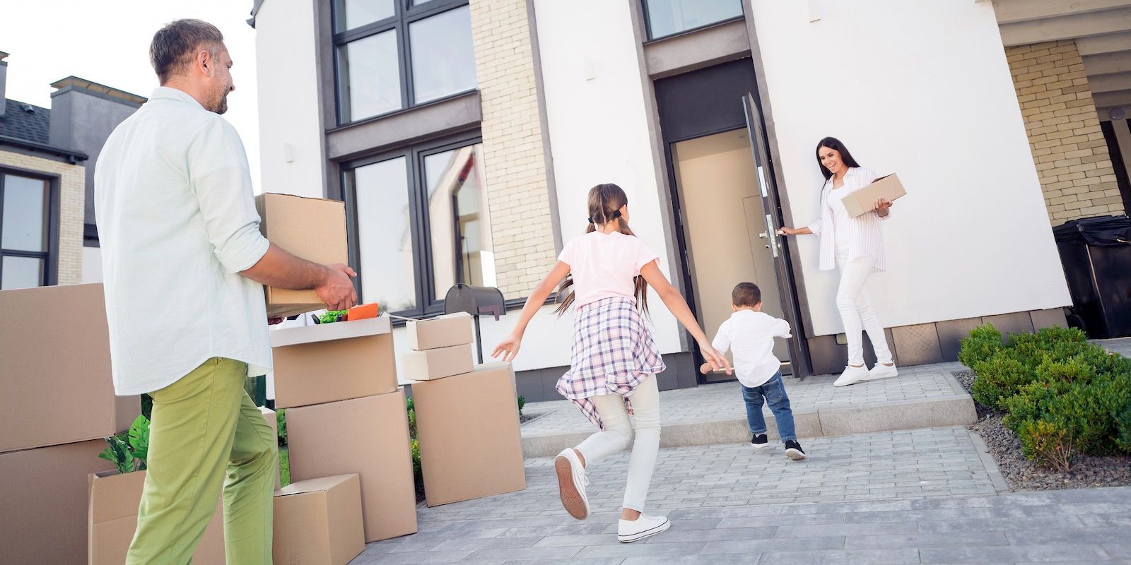 Family moving into a new house. Father carrying boxes, children running, mother holding a box at the open door.