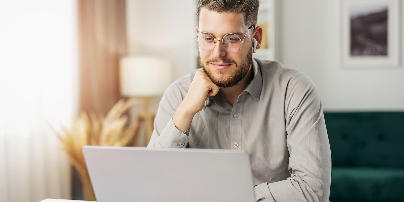 Man with glasses looking at a laptop, resting his hand on his chin, in a well-lit living room.