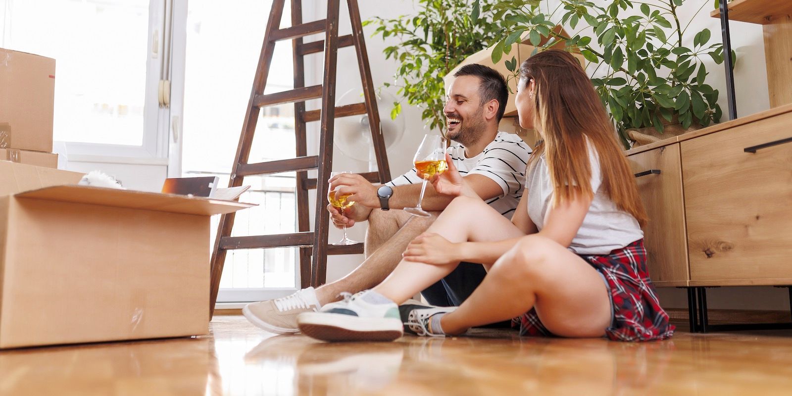 A couple sitting on a wooden floor, drinking, surrounded by moving boxes and furniture, smiling.