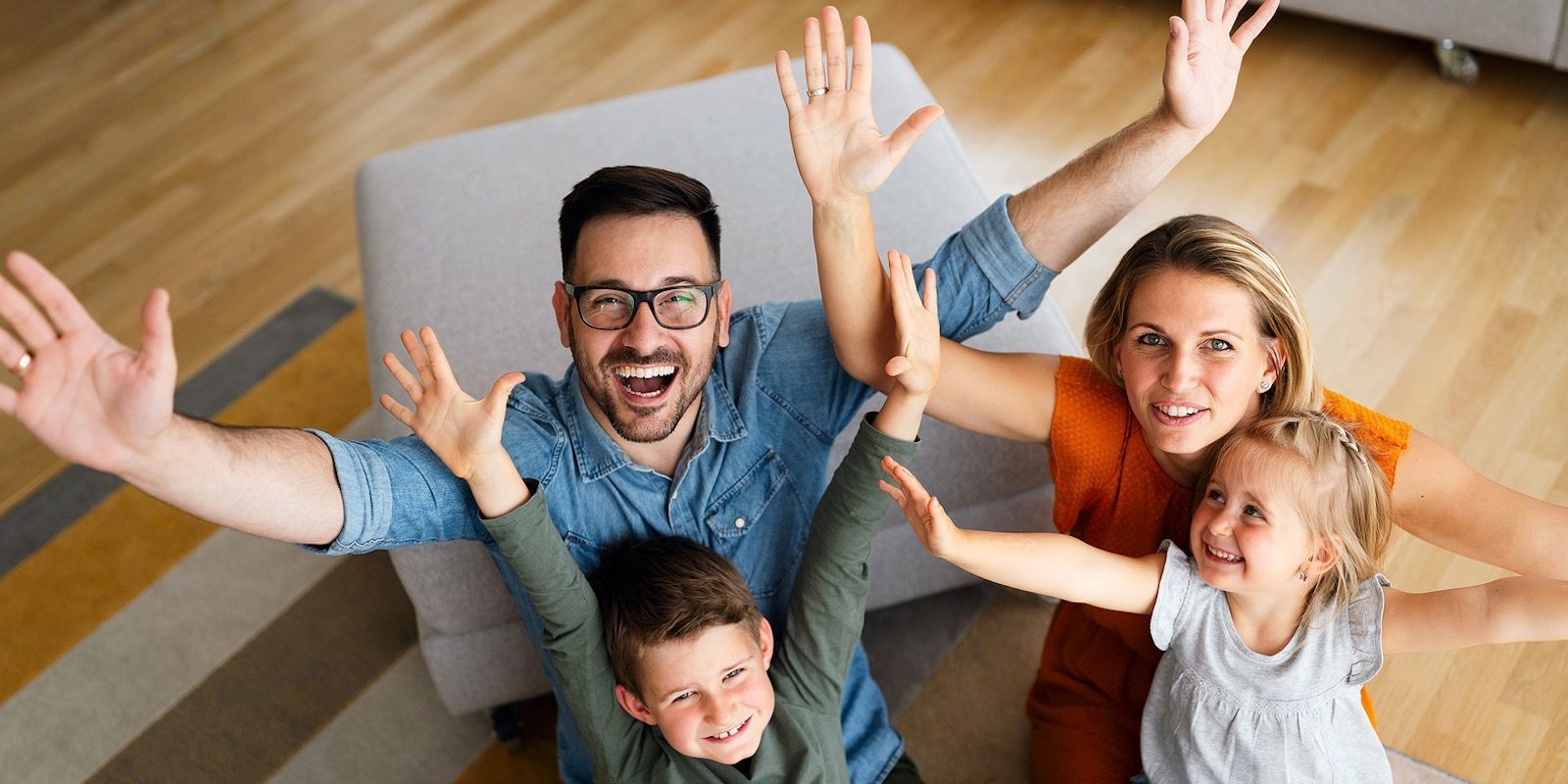 Family of four with arms raised, smiling at the camera. They are indoors on a wooden floor.