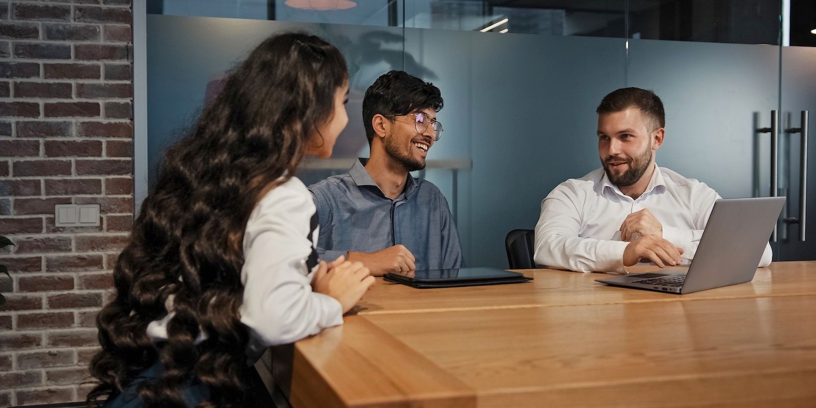 Three people at a table in an office. One person is using a laptop. They are talking and smiling.