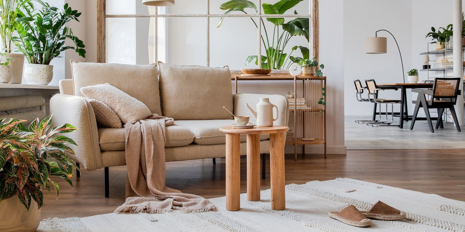 Cozy living room with beige sofa, wooden table, and many indoor plants. Dining area visible in the background.