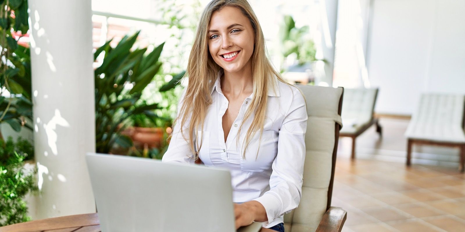 Woman with blonde hair wearing a white button-down shirt smiles while working on a laptop outdoors.