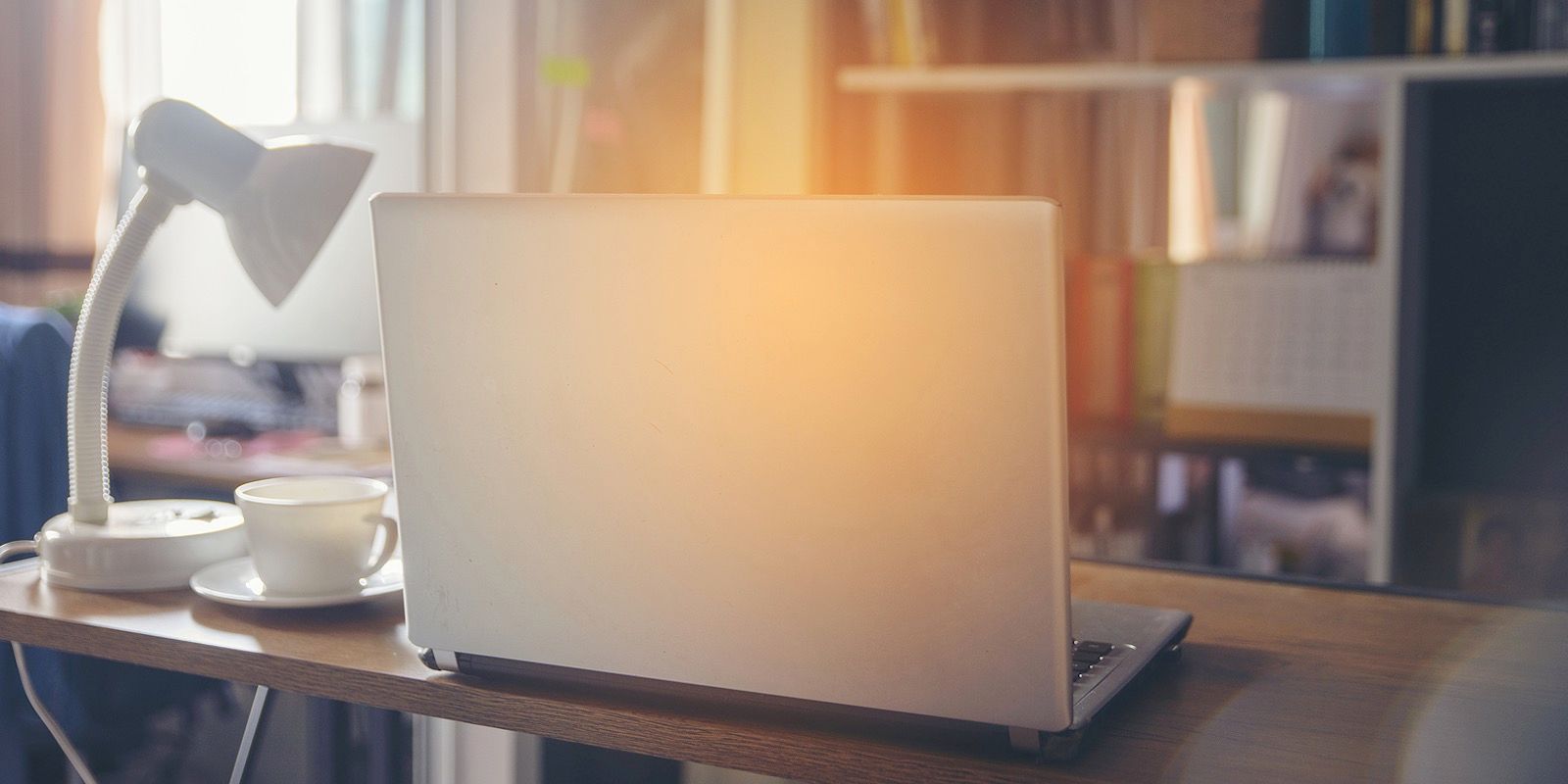 Laptop on a wooden desk with a lamp and a cup, in a home office setting, backlit by the sun.
