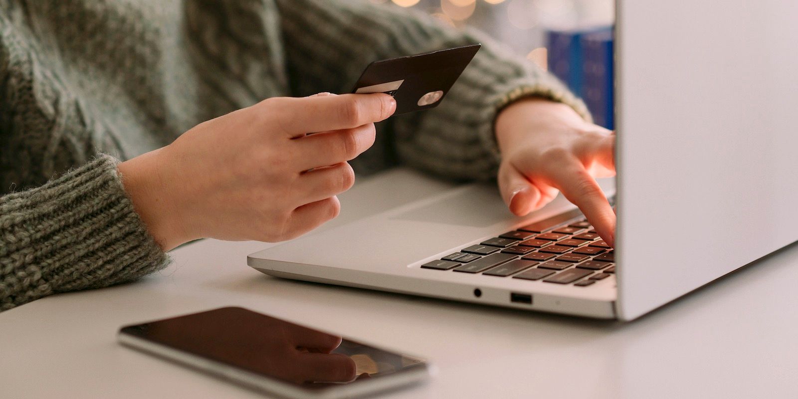 Person holds a credit card and types on a laptop. A smartphone lies on the table.