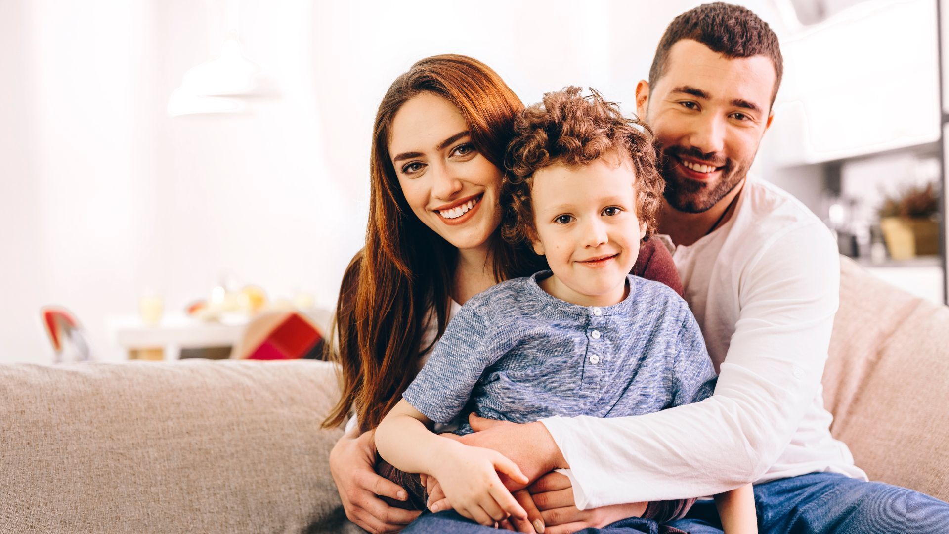 Family of three smiling on a couch; woman, man, and child; indoor, neutral tones.