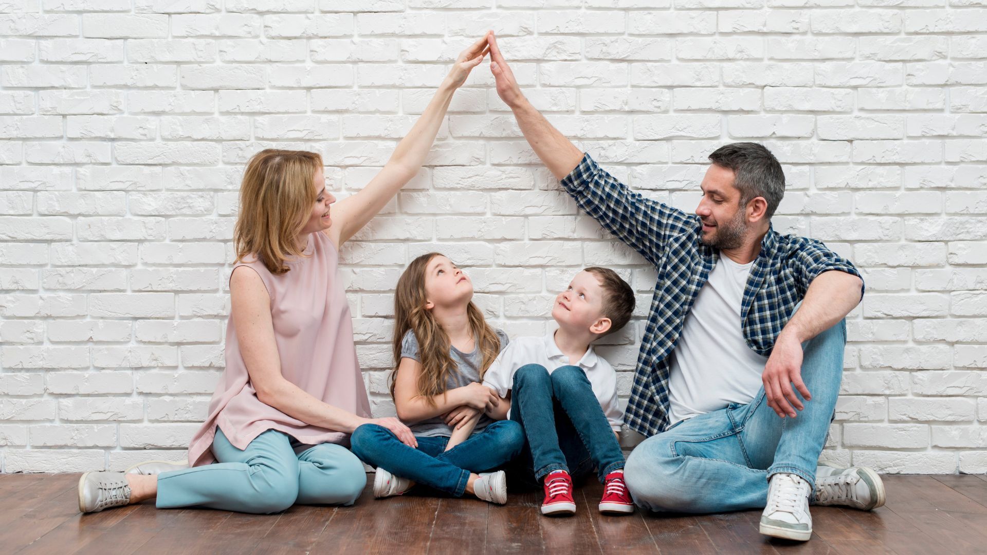 Family making a house shape with their arms in front of a white brick wall, smiling.