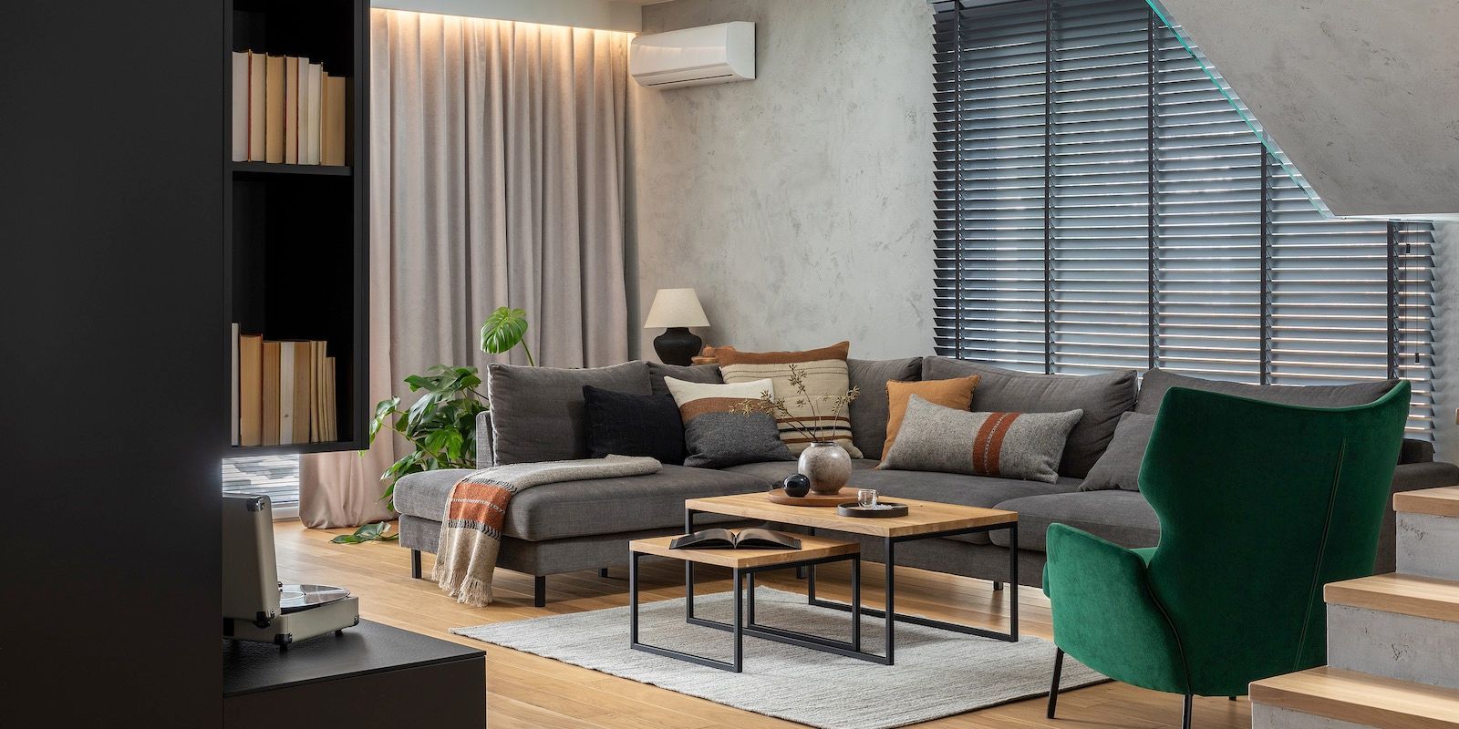 Living room with a dark gray sectional sofa, green armchair, and wooden coffee tables. Light curtains and a bookshelf on the left.