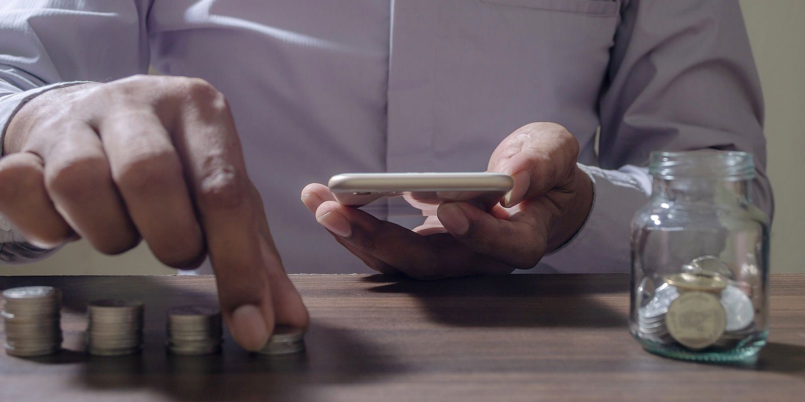 Person counting coins while holding a smartphone; jar of coins nearby.