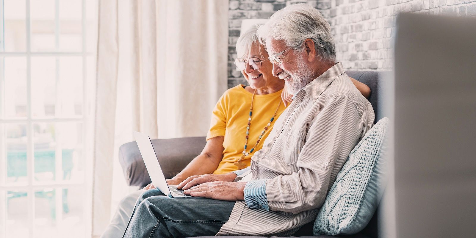 An elderly couple sits on a couch, looking at a laptop.
