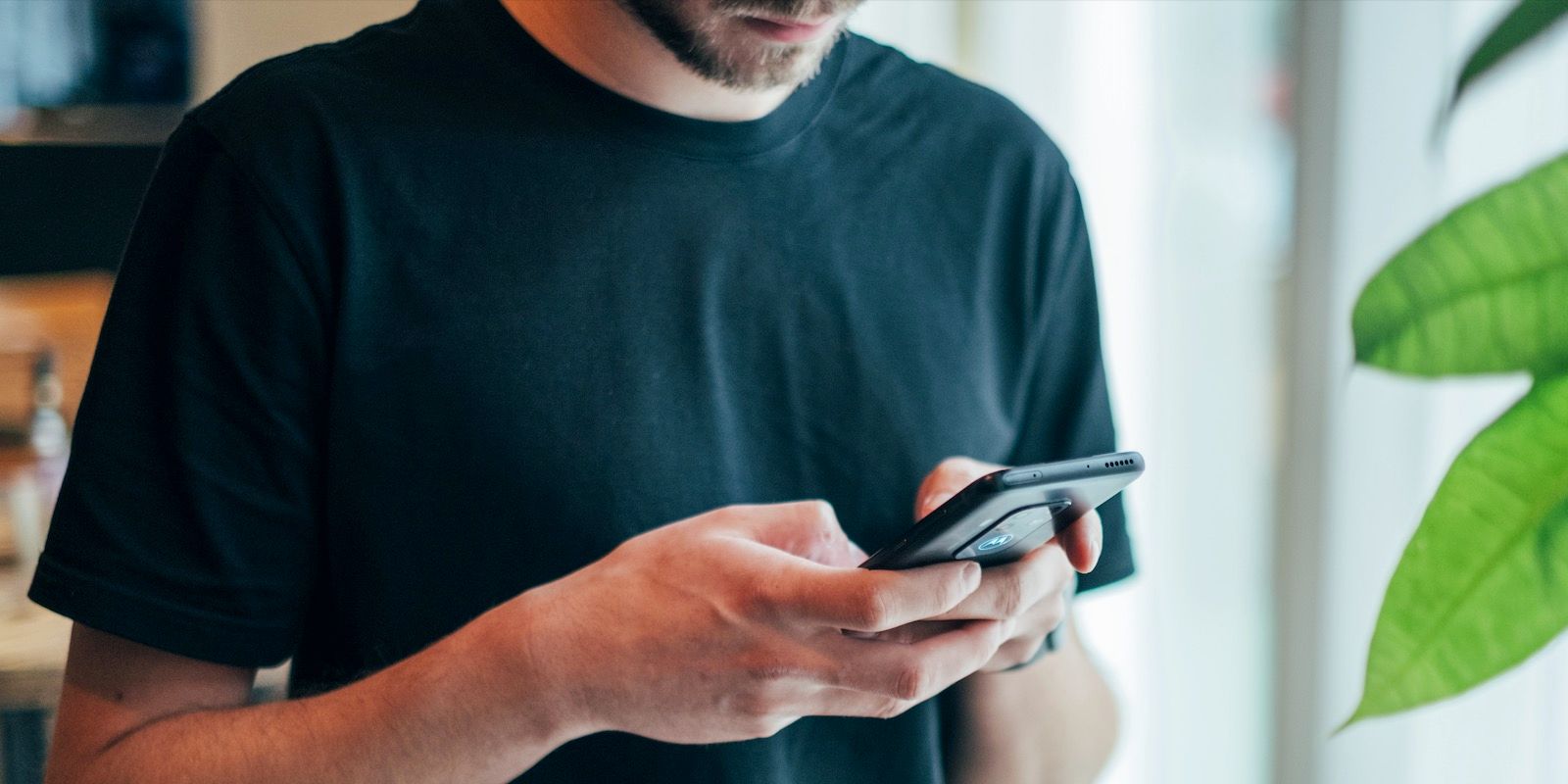 Person using a smartphone, holding it with both hands. Dark blue shirt, green plant in background.