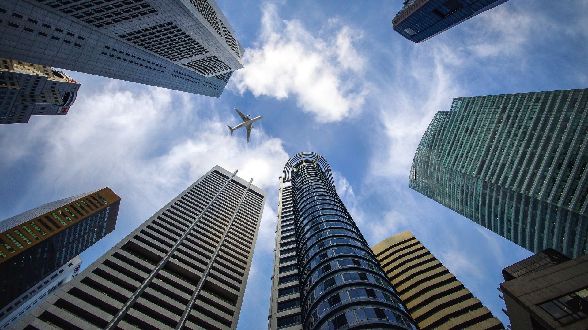 Skyscrapers viewed from below, airplane flying overhead against a cloudy sky.
