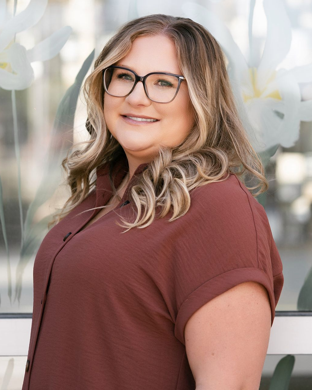 Woman in glasses, wearing a rust-colored shirt, smiling, posing in front of a window with white floral designs.