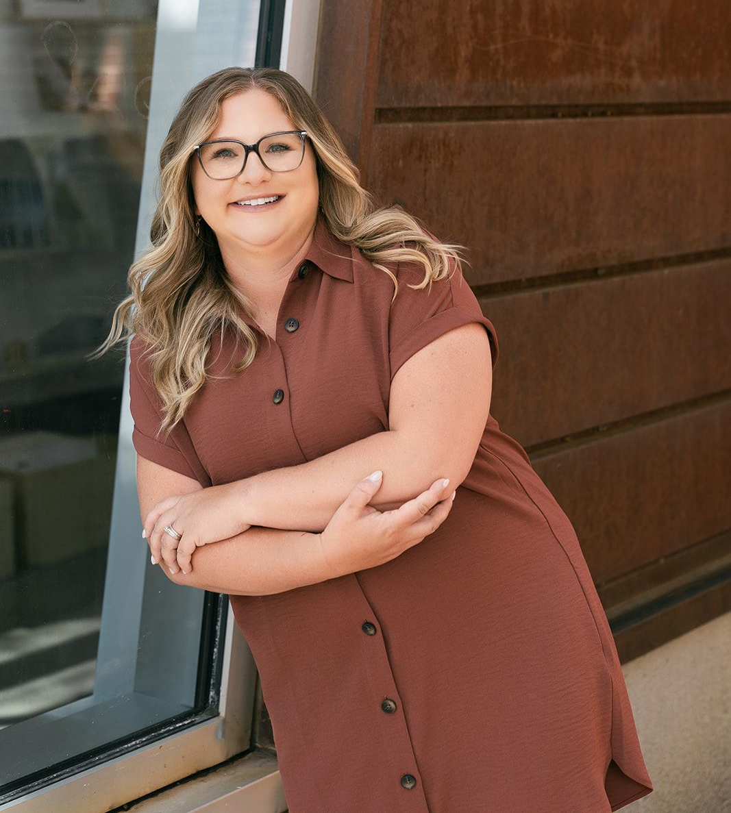 Woman in glasses and rust-colored dress, smiling, arms crossed, leaning against a building.