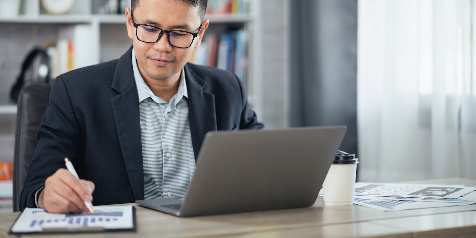 A person in glasses and a suit, working on a laptop and documents at a desk in an office.