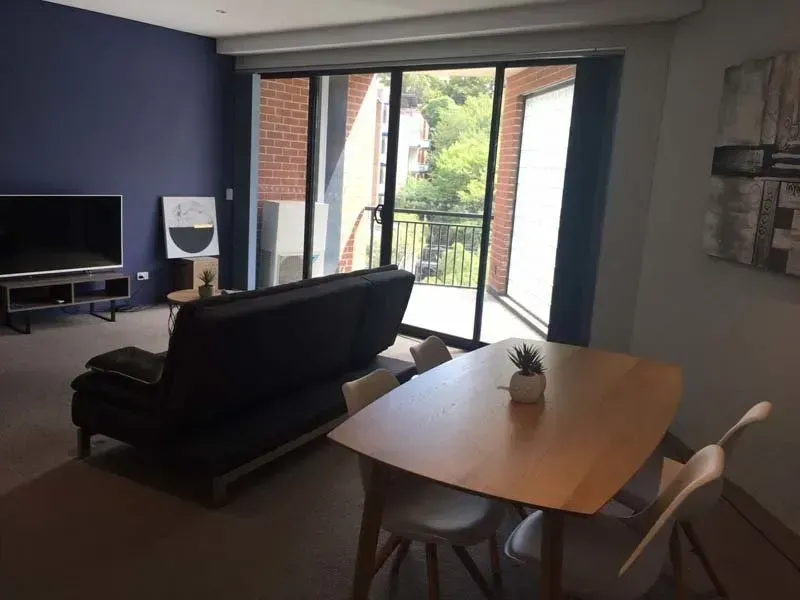 Living room with a black futon, wooden dining table, and balcony access. Blue wall, natural light.