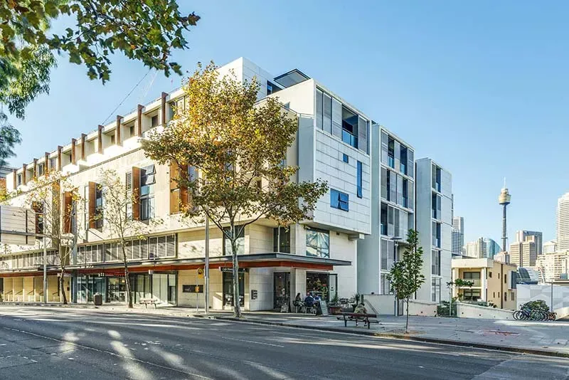 Street view of a modern multi-story building with shops below and apartments above. Tree in foreground, city skyline.