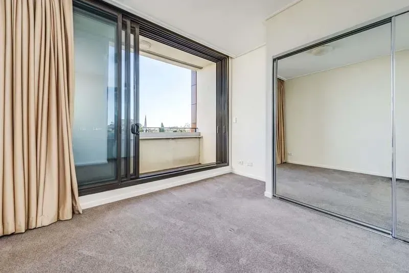 Empty bedroom with sliding glass door to balcony, mirrored closet doors, beige curtains, and gray carpet.