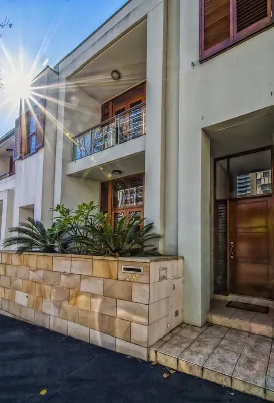 Cream-colored townhouse with wooden doors and shutters, a balcony, and a stone wall. Bright sunlight.