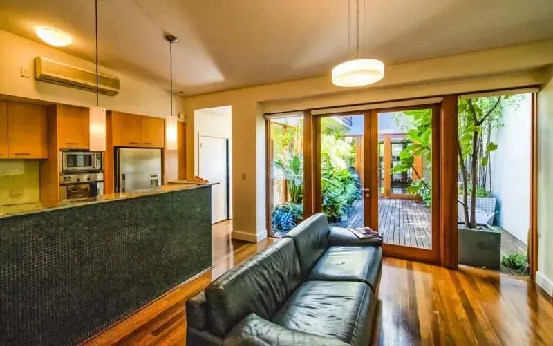 Living room with open wooden doors to a garden; black leather couch, wood flooring, kitchen with brown cabinets.