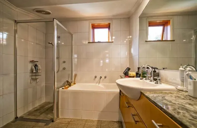 Bathroom with shower, tub, sink, and two small windows. White tile, wood vanity, and a large mirror.
