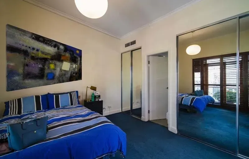 Bedroom with blue bed, large artwork, mirrored closet, and window with shutters.
