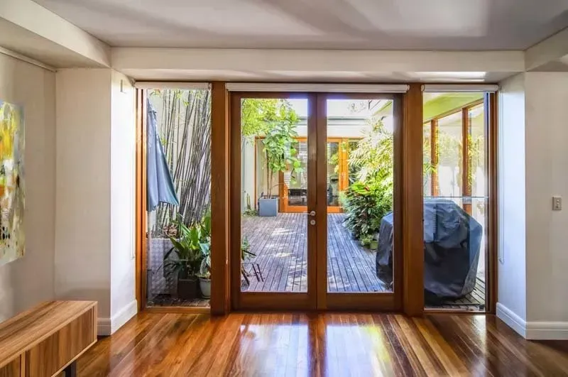 Wooden double doors open onto a patio. Lush greenery surrounds a covered grill and wooden deck.