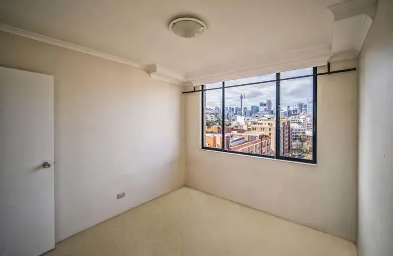 Empty, off-white room with a city view through a large window; a white door is on the left.