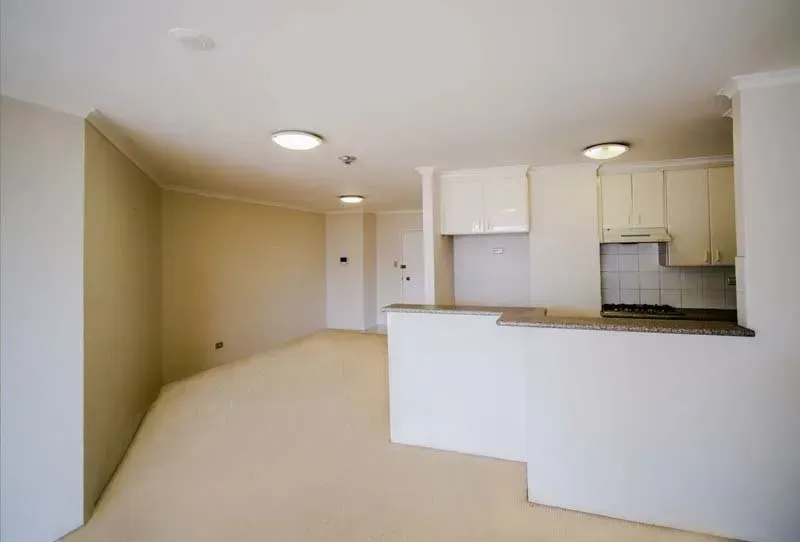 Empty apartment interior with kitchen and white walls, light beige floor, and recessed lighting.